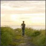 Mann von hinten läuft auf einem Küstenweg auf das Meer zu.