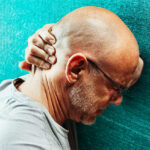 älterer Mann mit Glatze und weißem Stoppelbart hält sich den schmerzenden Hinterkopf